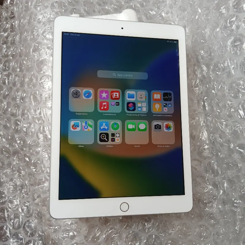 A white tablet with a black screen displaying various app icons is placed on a sheet of bubble wrap. The screen shows the "App Library" with categories such as "Recently Added," "Utilities," "Social," "Entertainment," "Productivity & Finance," "Other," and "Suggestions." The tablet has a home button at the bottom and a front camera at the top