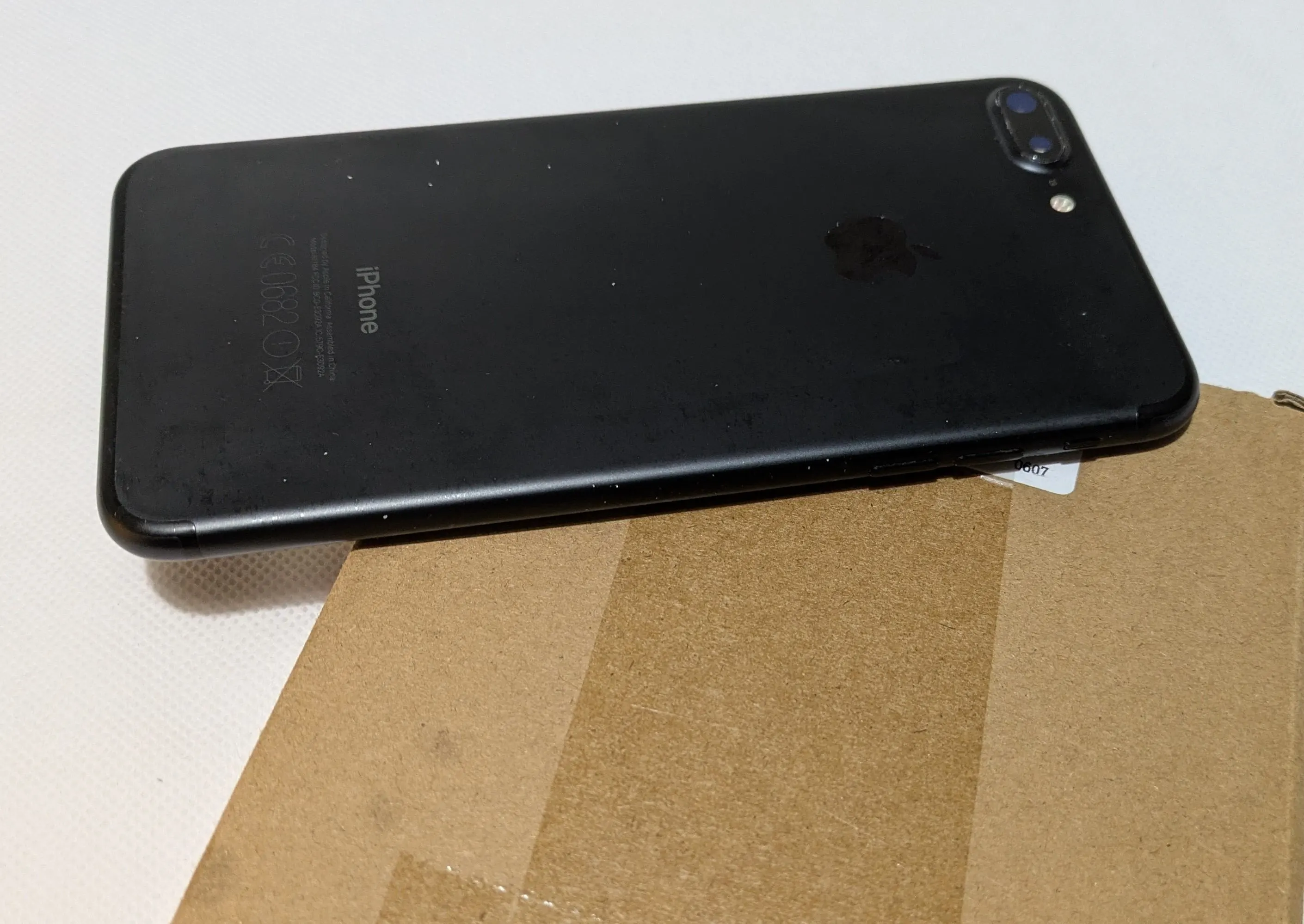 Back view of a black iPhone 7 Plus placed on a cardboard surface. The phone features a dual camera and flash at the top corner, along with the Apple logo in the center. Dust particles are visible on its surface, emphasizing its condition.