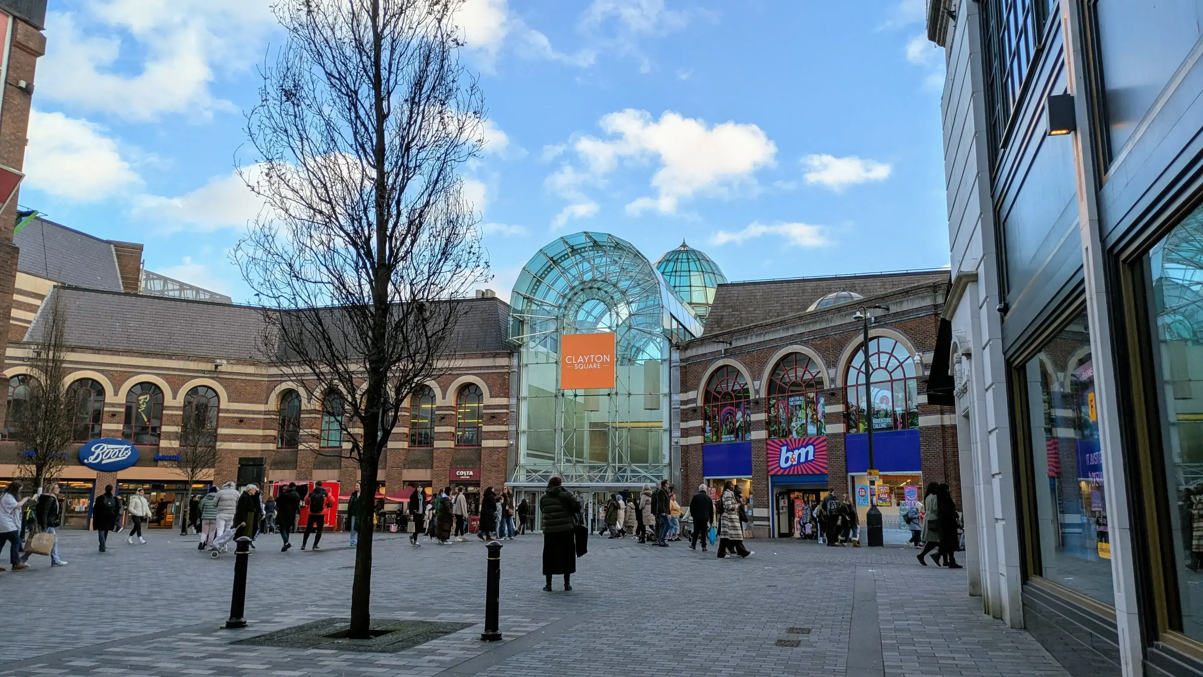 A view of Clayton Square Shopping Centre with people walking around the plaza. The image shows a glass entrance with a sign that reads "Clayton Square" and various shops, including Boots, Costa, and B&M. The architecture of the buildings is a mix of modern glass and traditional brickwork, making it an interesting blend of old and new styles. The sky is clear with a few clouds, suggesting a pleasant day.