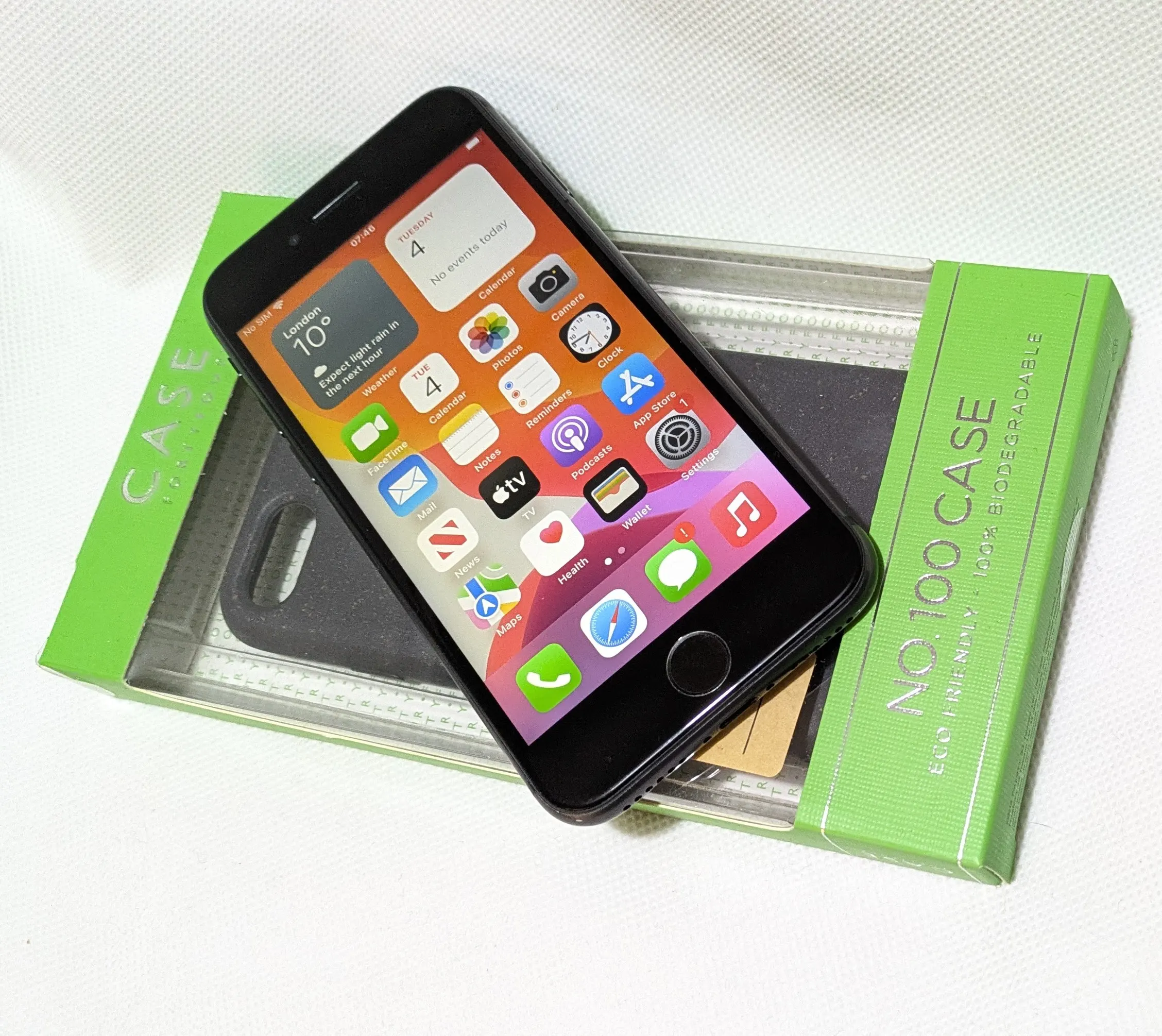A refurbished iPhone displaying the home screen with various app icons, placed on top of a green eco-friendly, biodegradable phone case packaging labeled 'NO.100 CASE' in Liverpool
