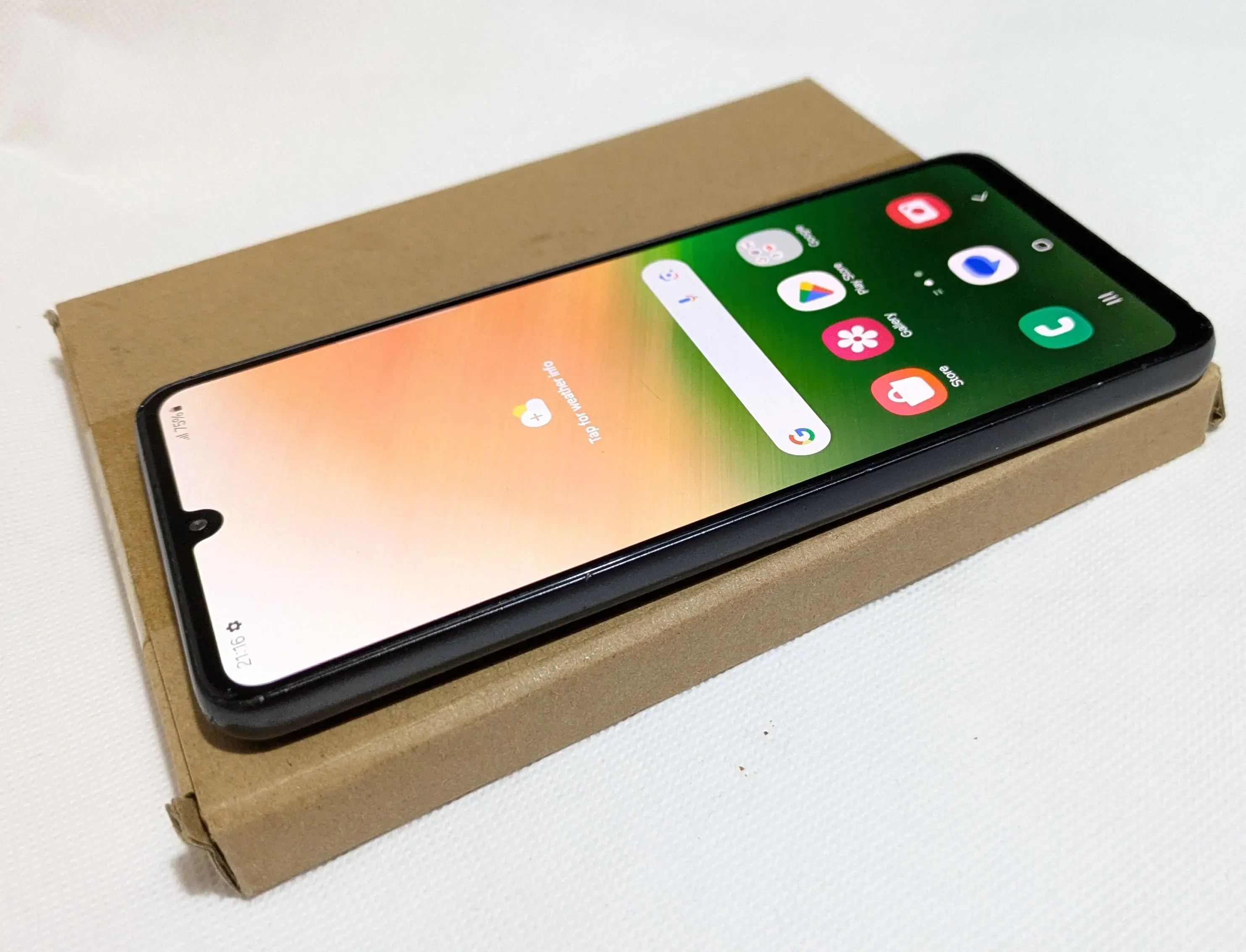 A sleek black smartphone displayed on a brown cardboard box, showcasing various app icons, including messaging, contacts, and the Google Play Store. The screen background features a vibrant gradient of orange and green
