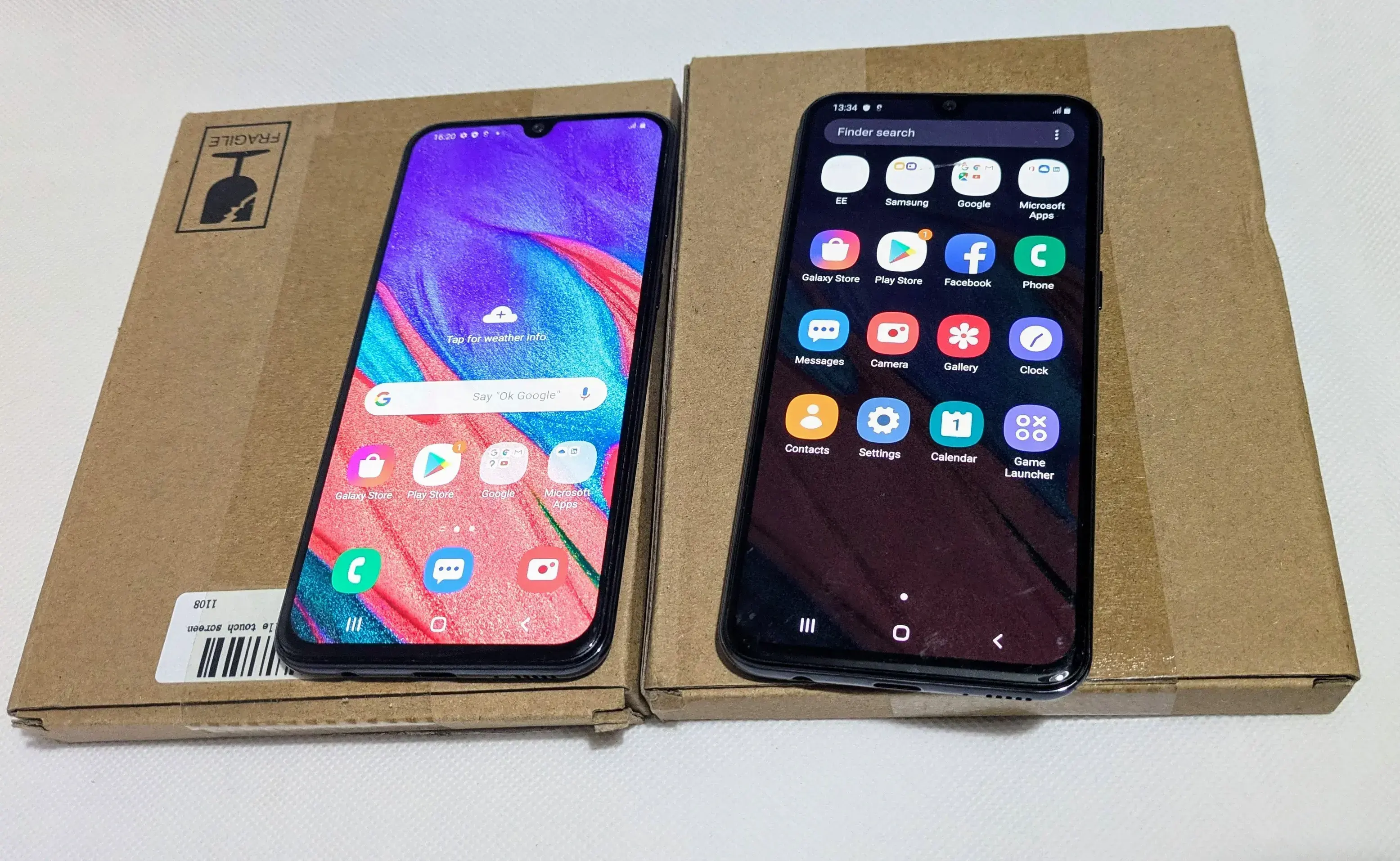Two Samsung Galaxy A40 smartphones placed on cardboard boxes. The phone on the left displays the home screen with various app icons and a colorful wallpaper, while the phone on the right shows the app drawer with multiple app icons.