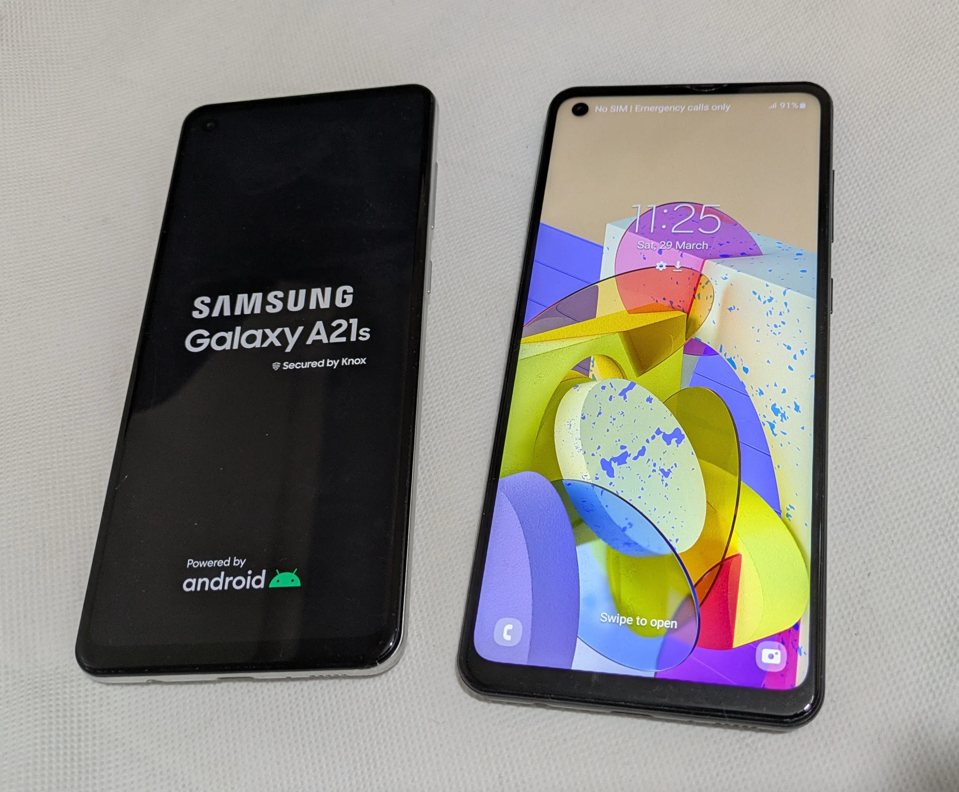 Two Samsung Galaxy A21s smartphones for sale in Liverpool, displayed on a light surface. One phone shows the startup screen with 'Samsung Galaxy A21s Secured by Knox Powered by Android,' while the other displays the home screen with colorful abstract wallpaper and system details