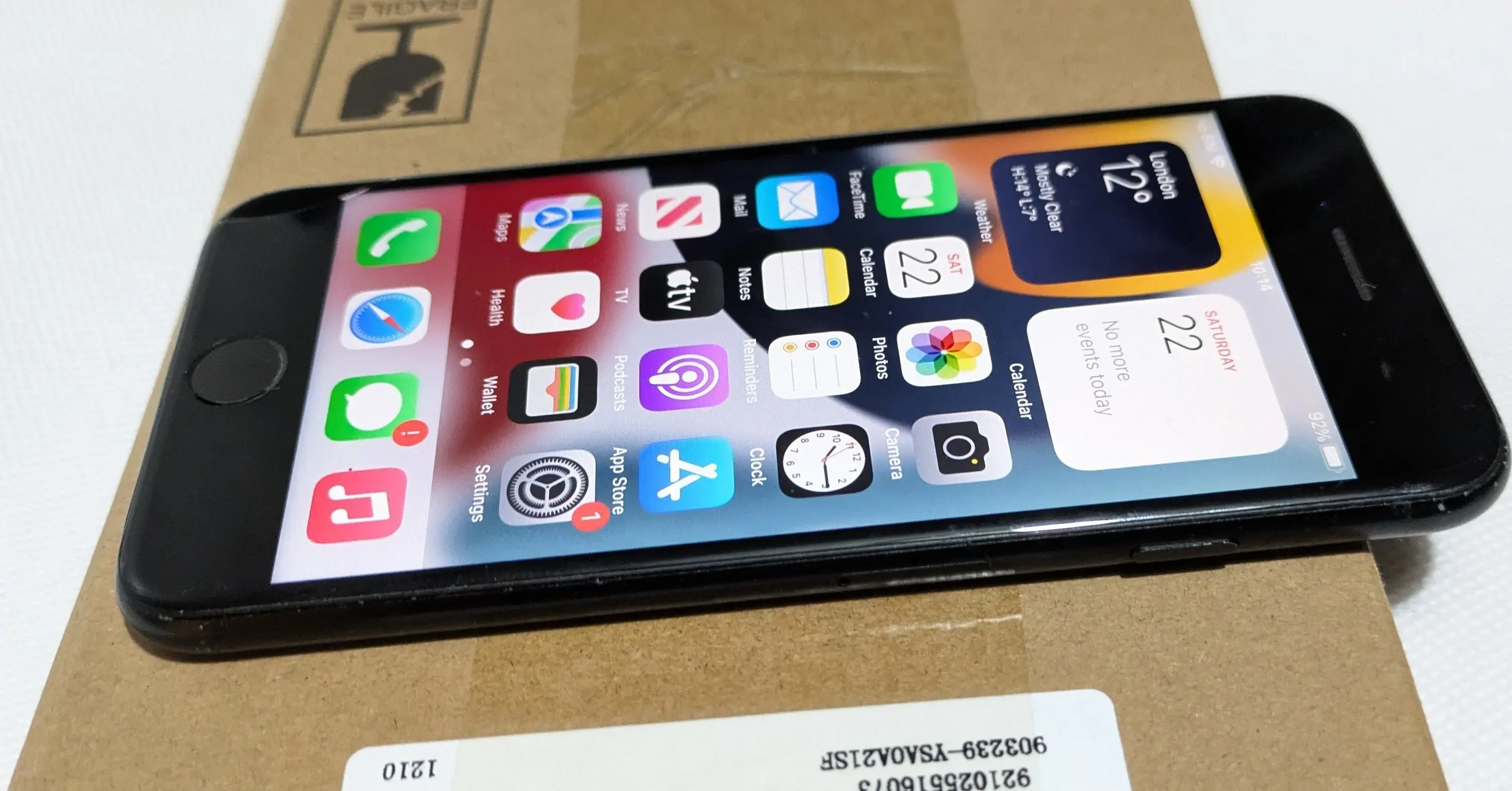 A black smartphone with various app icons displayed on the screen, resting on a cardboard box. The screen shows the date as Saturday, 22nd, and the weather in London as mostly clear with a temperature of 12 degrees Celsius.