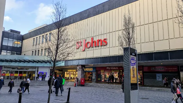 The image shows the exterior of St Johns Shopping Centre in Liverpool. The building has a large sign with "St Johns" in red letters. Below the sign, there are several shops, including McDonald's, Warren James, Greggs, Ryman, and Shush. People are walking in the pedestrian area in front of the shopping centre. There is also a tall advertisement board in the foreground. The sky is partly cloudy.