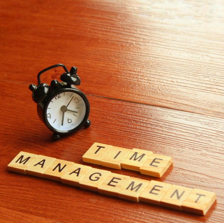 Analog alarm clock beside wooden tiles spelling ‘Time Management’ on a wooden surface.